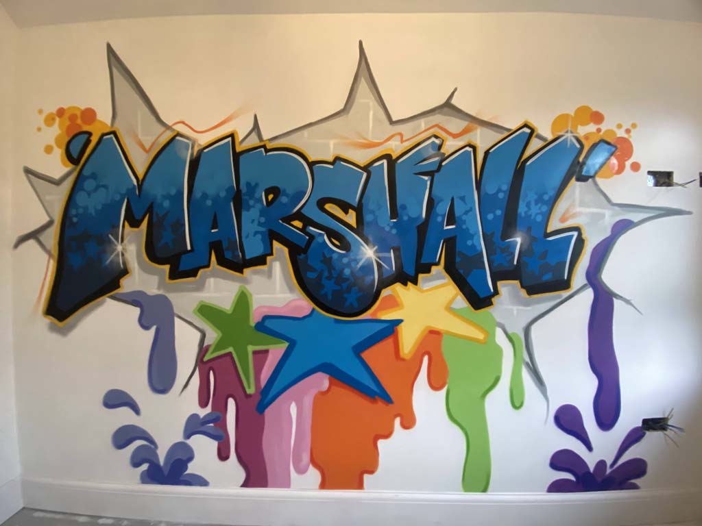 A bedroom wall spraypainted with the name Marshall in a graffiti style in blues with stars and splashes of bright colours below