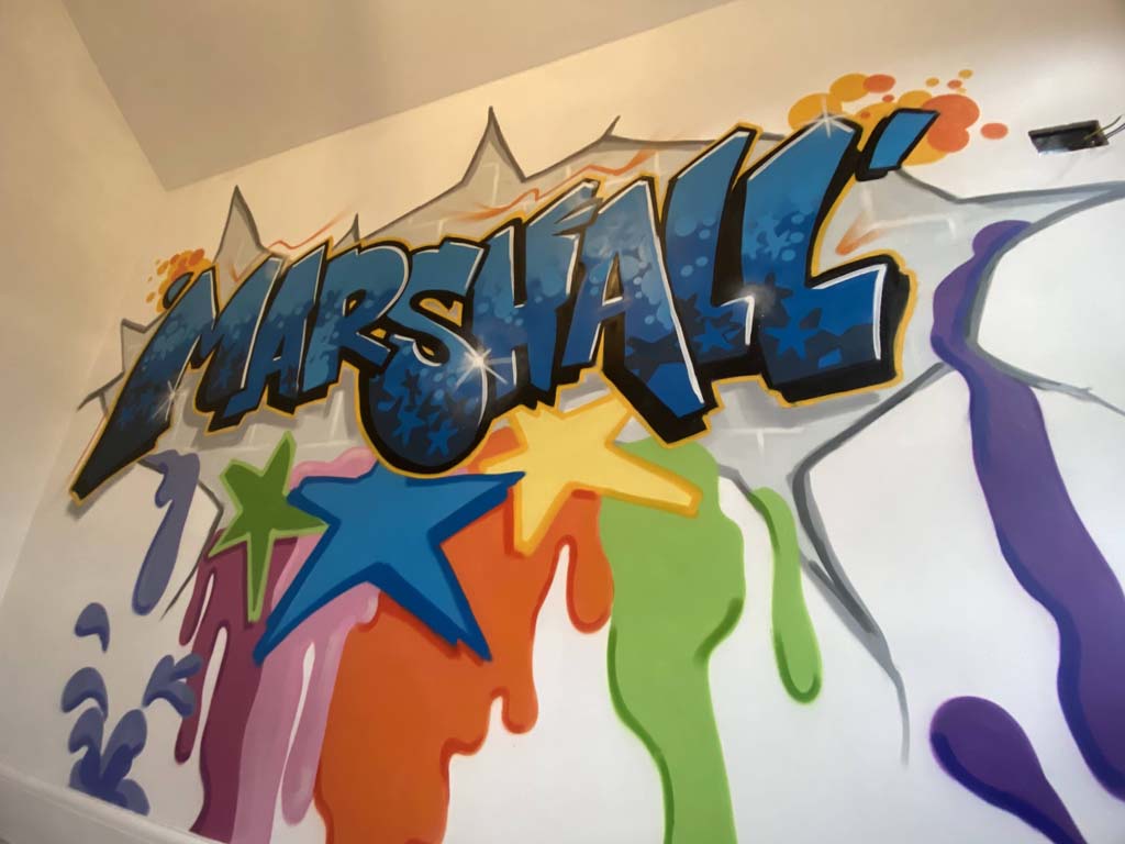 A bedroom wall spraypainted with the name Marshall in a graffiti style in blues with stars and splashes of bright colours below